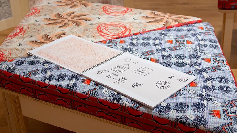 A sketchbook with line drawings is open on a wooden bench with brightly coloured and patterned cushions